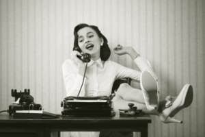 a sepia toned picture of a woman on an old phone with her legs up on the desk and a typewriting in front of her
