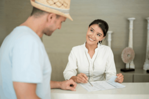 a woman employee smiling and showing a man in a hat something on a piece of paper from behind a hotel desk