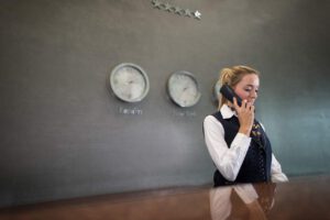 woman holding a phone while looking down and standing behind a desk. Clocks with the time of London and New York and four stars are on the gray wall behind