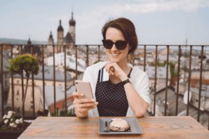 a woman in sunglasses wearing a black dress with polka dots smiling at her smartphone with a payment card in her hand sitting on a rooftop restaurant