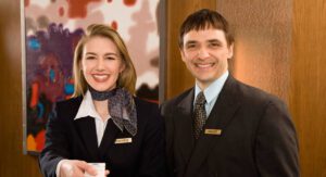 a woman wearing a black suit and neck scarf holding out a hotel key next to a hotel manager in a suit, both smiling and looking at the camera