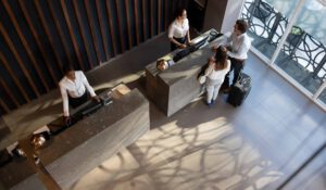 arial photo of two people checking into a hotel with two employees behind the desk with stone desks, a window showing the street outside and wood accents on the wall