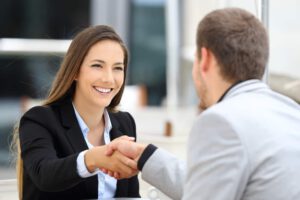 woman smiling and wearing a black suit and blue top shaking hands with a man in a grey suit