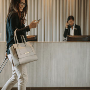 a woman smiling at her smartphone while rolling a suitcase through a hotel lobby with a hotel employee behind a desk in the background