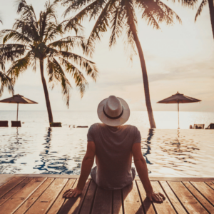 person in a hat facing away from the camera, feet dangling into an infinity pool with an ocean and palm trees in the background