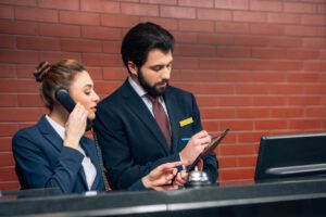 two hotel employees wearing suits with one on the phone and one writing things down behind a hotel desk with a brick wall in the background