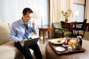 a man wearing a blue shirt drinking coffee and using a laptop while sitting on a couch near an ottoman with breakfast sitting on it in a hotel room