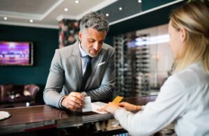 a man with gray hair and a light gray suit receiving paper from a hotel concierge over a hotel desk