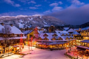 a ski resort with snow on the rooftops, outdoor lights on, snow on the ground, and ski hills in the background with mountains and pine trees