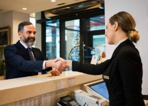 a man in a suit and graying beard shaking hands with a female concierge from behind a hotel desk