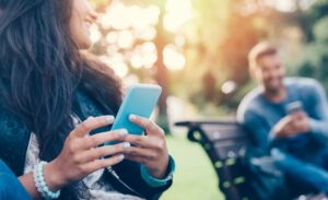 close up of a woman holding a smart phone while sitting on a park bench and a man using a smartphone in the background