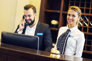 a man in a suit on the phone and a woman next to him smiling, both behind a hotel front desk