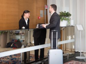 a man with a small grey suitcase at a hotel concierge desk talking to a woman behind the desk