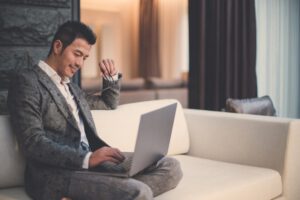 man in a gray suit sitting on a white couch looking at a computer while in a hotel