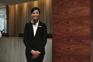 woman wearing a black suit, smiling and facing the camera in front of a hotel concierge desk and wood wall