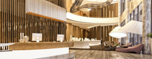 two story hotel lobby with large lighting, a stone wall, front desk with wood accents and two computer monitors, and a large hanging ceiling decoration. tones of brown, yellow, white, grey and gold
