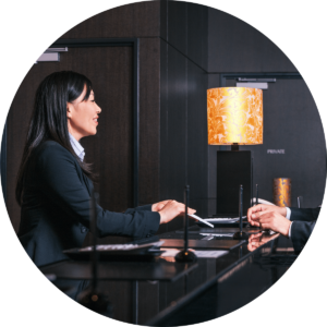 woman wearing a suit standing behind the front desk of a hotel and handing someone a piece of paper, dark wood walls and a orange decorative lampshade