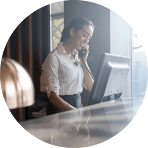 A woman wearing a white top, smiling while on the phone and looking at a computer monitor at the front desk of a hotel