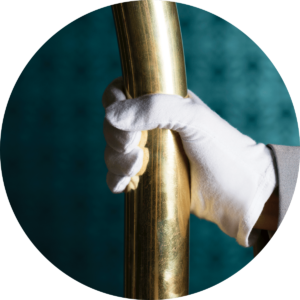 A hand wearing a white glove holding onto a gold pole of a luggage cart with a turquoise wall behind